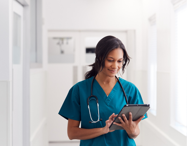 Female Doctor Wearing Scrubs In Hospital Corridor Using Digital Tablet