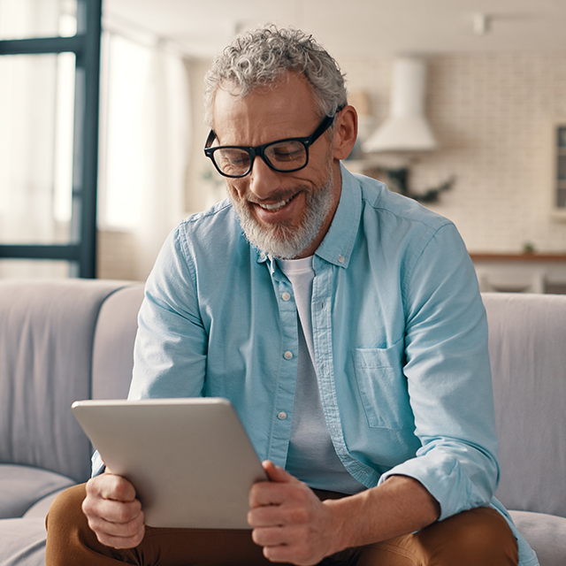 Nest Pension Services images - Cheerful senior man in casual clothing using digital tablet while sitting on the sofa at home